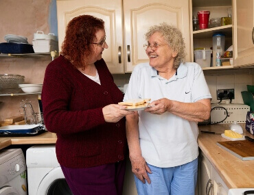 elderly companion and elderly lady handing a plate to the other